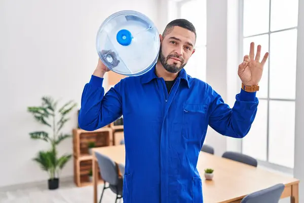 Hispanic service man holding a gallon bottle of water for delivery doing ok sign with fingers, smiling friendly gesturing excellent symbol