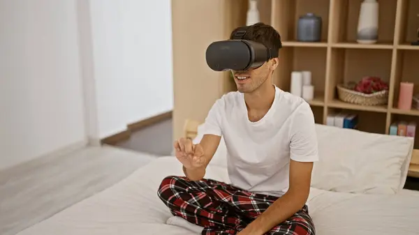 Smiling young hispanic gamer guy enjoying sitting in pyjama on bedroom bed, playing video game using futuristic vr glasses