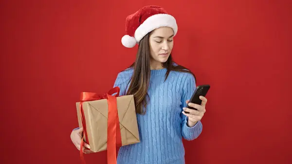 年轻的高加索女人戴着圣诞礼帽 用智能手机把礼物放在孤立的红色背景上 — 图库照片
