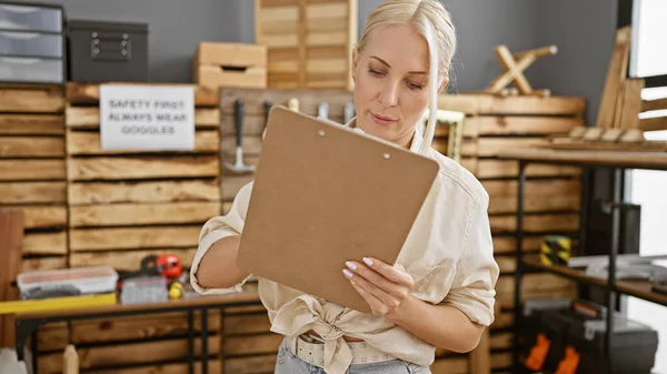 Attractive young blonde woman carpenter takes diligent notes on clipboard during woodwork in bustling carpentry workshop
