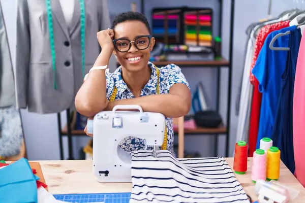 African american woman tailor smiling confident leaning on sewing machine at tailor shop