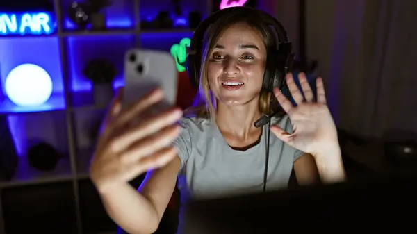 Smiling young blonde streamer woman enjoying the game, streaming live video call from her gaming room at home, engrossed in her virtual world of digital entertainment