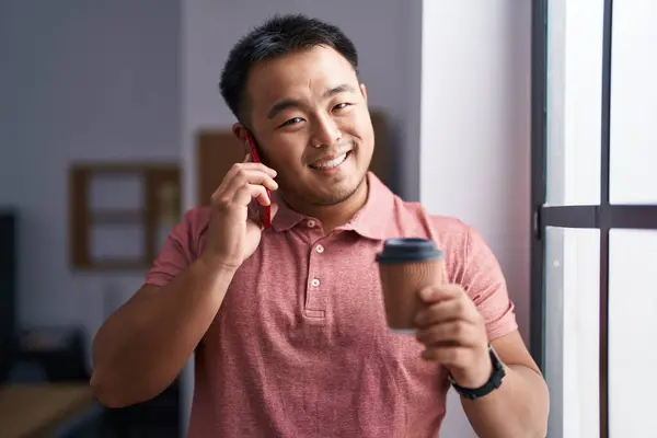 Young chinese man business worker talking on smartphone drinking coffee at office