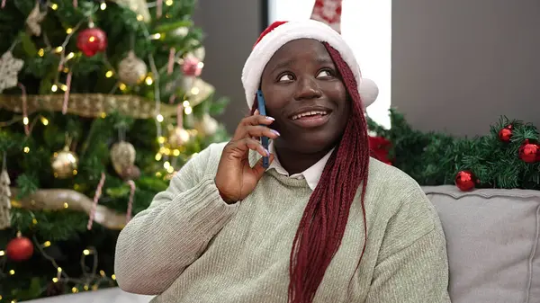 African woman with braided hair speaking on the phone by christmas tree at home