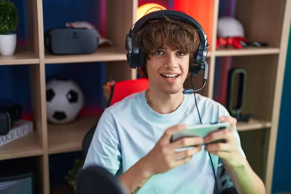 Young blond man streamer playing video game using smartphone at gaming room