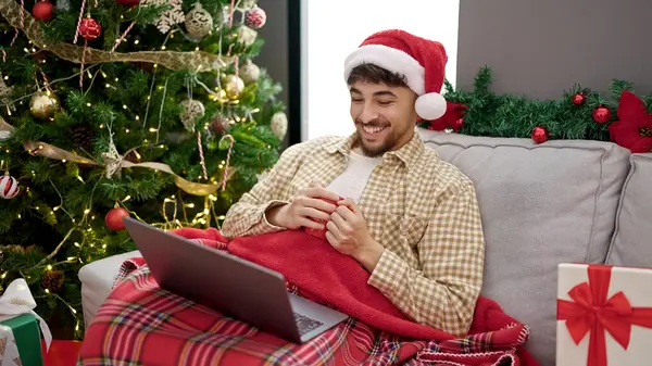 Young arab man watching movie drinking coffee sitting by christmas tree at home