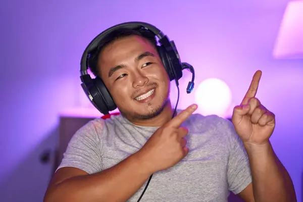 中国の若い男はヘッドフォンを笑顔で身に着けているビデオゲームをプレイし 側面に2つの手と指を指してカメラを見ています — ストック写真