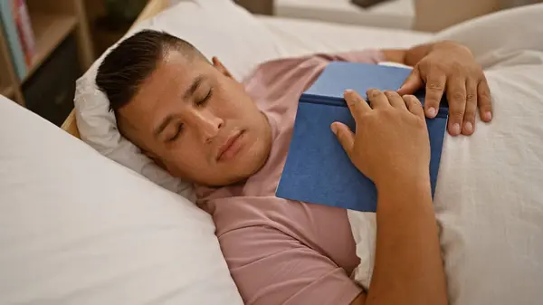 Handsome young latin man, cozily sleeping away the morning, book comfortably resting on his chest, lying relaxed in the comfort of his bedroom bed at home