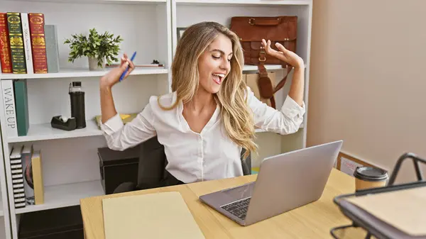 Confident young woman dances to the beat of success at work. blonde business professional celebrates with joy, laptop background on the office desk