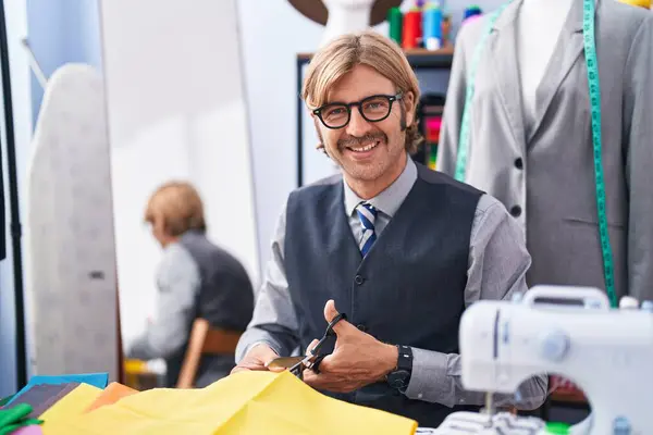 Young blond man tailor smiling confident cutting cloth at clothing factory