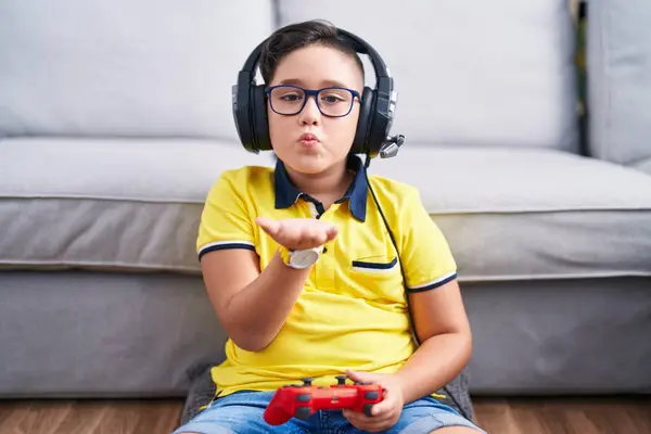 Young hispanic kid playing video game holding controller wearing headphones looking at the camera blowing a kiss with hand on air being lovely and sexy. love expression.
