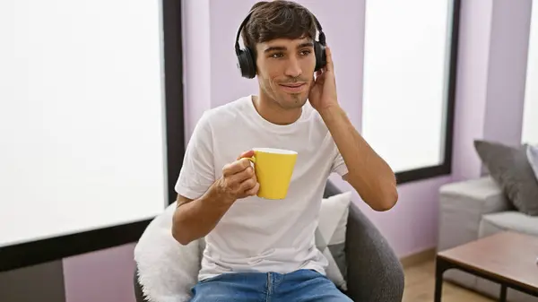 Young, handsome hispanic man joyfully enjoying his morning espresso on the sofa, at home, totally relaxed while sinking in the sounds of his favorite music through his headphones.