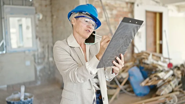 Middle age blonde woman architect talking on smartphone writing on document at construction site