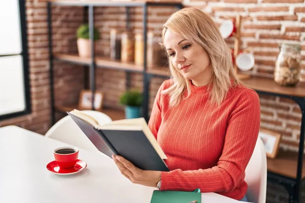 Young blonde woman reading book and drinking coffee at home