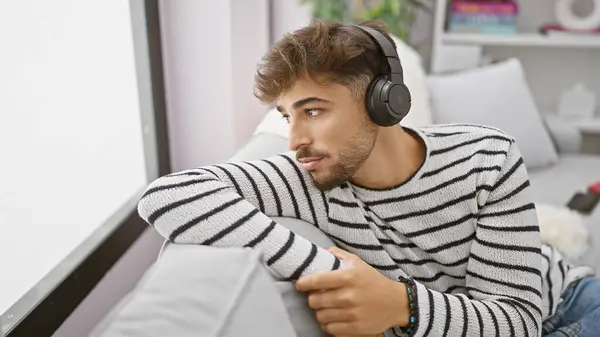 年轻的阿拉伯男子喜欢自己的声音避难所 在沙发上放松 全神贯注于耳机的音响技术 在家里听音乐 — 图库照片