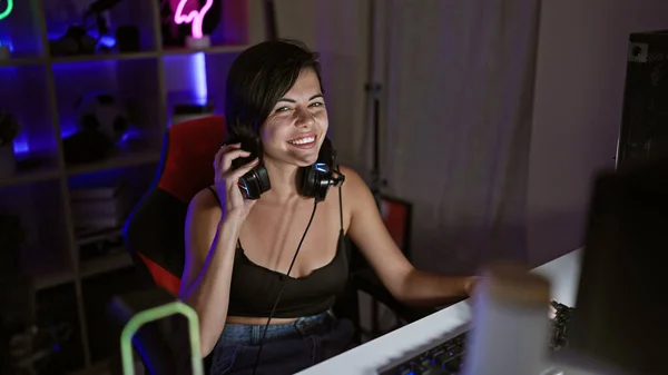 Vibrant young hispanic woman streamer confidently smiling, sporting headphones in her atmospheric gaming room, immersed in a futuristic cyber-stream gaming experience