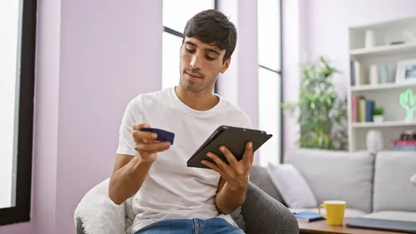 Tech-savvy young hispanic man relaxing on sofa, shopping online with credit card and touchpad indoors - portrait of serious but relaxed shopper engaging in digital retail from the comfort of home