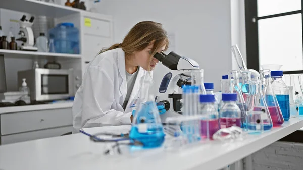 科学の中心では 若い魅力的なブロンド女性科学者が意図的に研究室で顕微鏡で作業し 分析によって発見を醸成 — ストック写真