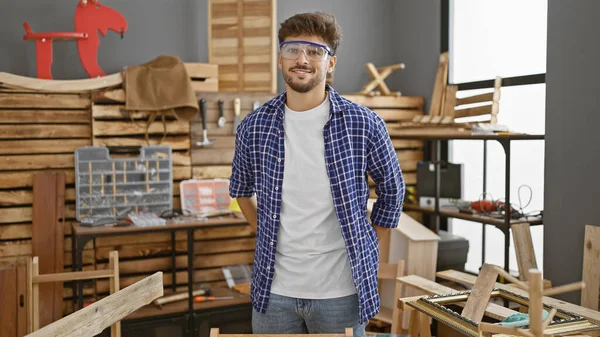 Smiling young arab carpenter, glasses donned, confidently standing in carpentry workshop, enjoying his woodworking profession