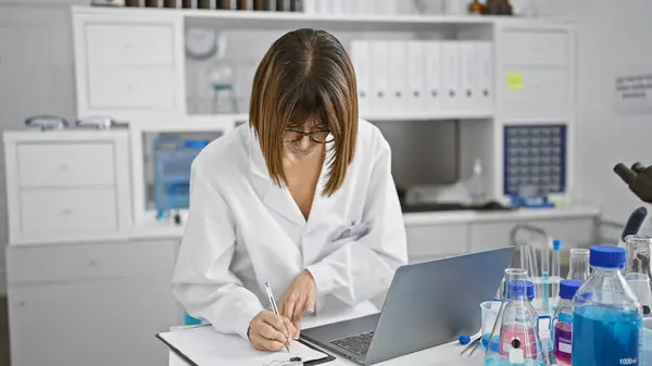 In the heart of science, a young, beautiful hispanic woman scientist sitting in a laboratory, engrossed in her medical research. writing notes, using a laptop, she discovers new frontiers.