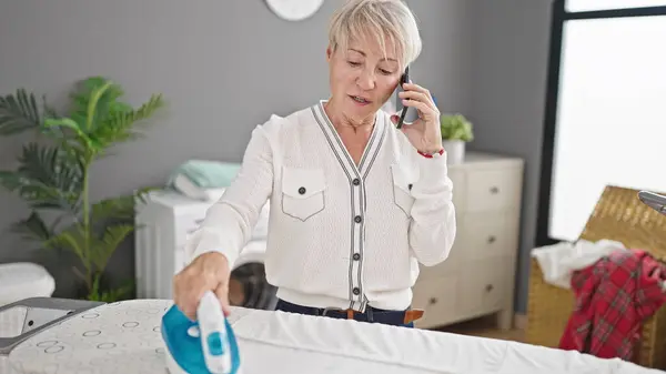 Middle age blonde woman talking on smartphone ironing clothes at laundry room