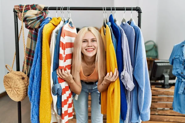 Young blonde woman customer smiling confident between clothes at clothing store