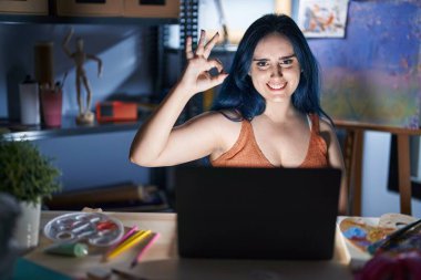 Mavi saçlı genç ve modern bir kız geceleri dizüstü bilgisayarla resim stüdyosunda oturuyor, gülümsüyor ve el ve parmaklarıyla 