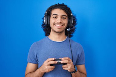 İspanyol asıllı, kıvırcık saçlı, video oyunu oynayan, elinde kumandayla gülümseyen, yüzünde mutlu ve havalı bir gülümsemeyle. diş gösterme. 