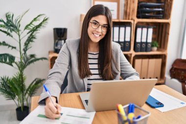 Genç, güzel İspanyol iş kadını. Ofisteki evraklarda dizüstü bilgisayar kullanıyor.