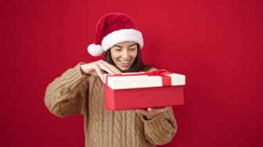 Genç, güzel, İspanyol bir kadın Noel şapkası takıyor, hediyesini açıyor ve izole edilmiş kırmızı arka planda üzgün görünüyor.