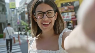 Tokyo şehri güzel İspanyol kadın olarak ışıldıyor, gözlük takıyor, eğlenceli bir video görüşmesine katılıyor. Gülümsemesi, kendine güveni şehir sokaklarında mutluluk saçıyor..