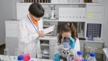 Mikroskop altında birlikte çalışan iki ciddi bilim kızı laboratuvar odasında özenle notlar alıyorlar. Tıbbi analiz ve araştırma alanında profesyonel deney.