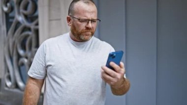 Neşeli, gözlüklü, sakallı, orta yaşlı, beyaz bir adam canlı, şehir sokağında telefonundan bangır bangır müzik dinliyor