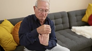 Beyaz saçlı, ağrıyan eli olan, yıpranmış bir adam, ciddi bir görünüm sergiliyor, odasındaki koltukta dinlenirken, evde artritten muzdarip.