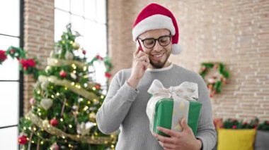 Noel şapkalı İspanyol adam elinde hediyeyle evde telefonla konuşuyor.