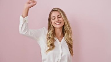 Neşeli genç sarışın kadın, gündelik gömlekli, güvenle müzik eşliğinde dans eden, neşe ve neşe dolu, pembe arka planda soyutlanmış, büyüleyici bir gülümsemeyle ışıldayan