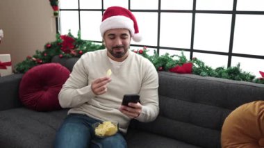 Genç İspanyol adam akıllı telefon kullanıyor ve cips yiyor. Evde Noel ağacının yanında oturmuş patates yiyor.