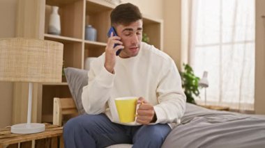 Hafta sonu uyanıyor, yakışıklı İspanyol genç adam yatakta dinleniyor, sabah kahvesinin ve yatak odasının tadını akıllı telefondan çıkarıyor.