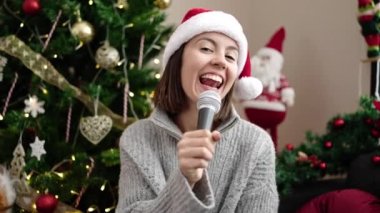 Genç ve güzel İspanyol kadın evdeki Noel ağacının yanında kanepede şarkı söylüyor.