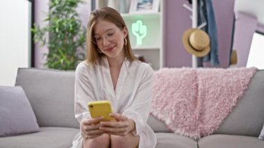 Kanepede oturmuş akıllı telefon kullanan genç sarışın kadın evde gülümsüyor.