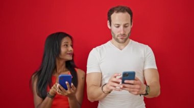Güzel çift, izole edilmiş kırmızı arka planda gülümseyen akıllı telefonlar kullanıyor.