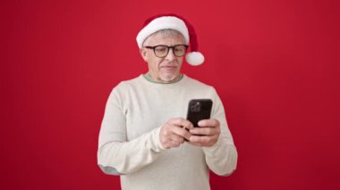 Orta yaşlı, gri saçlı, Noel şapkası takan, izole edilmiş kırmızı arka plan üzerinde parmağıyla akıllı telefonu işaret eden bir adam.