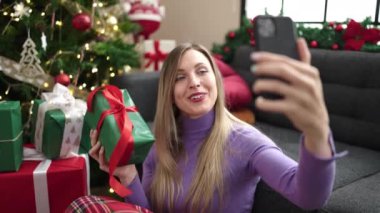 Genç sarışın kadın, evdeki Noel ağacının yanında oturmuş video görüşmesi yapıyor.