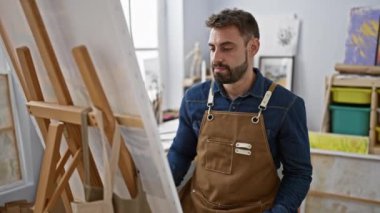 Sakallı genç bir İspanyol, kendine güvenen bir sanatçı, bir sanat stüdyosundaki tuvale tutkuyla çizerken gülümseyen ve terleyen.