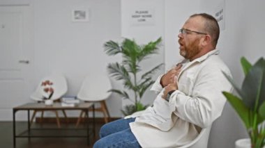 Yakışıklı, orta yaşlı, sakallı ve gözlüklü, mutsuz bir şekilde sandalyede oturan, korku dolu 19 korkunun ortasında bekleme odasında öksüren beyaz bir adam. Kapalı alandaki stresin üzgün portresi..