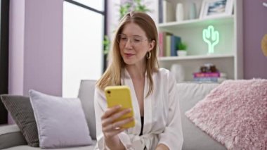 Genç sarışın kadın akıllı telefonu işaret ediyor. Kanepede oturuyor ve gülümsüyor.