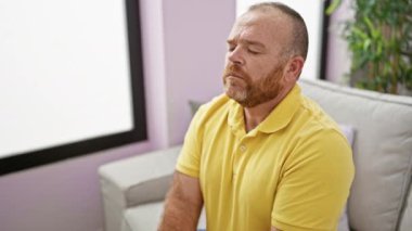 Yakışıklı orta yaşlı beyaz adam rahatça dinleniyor, meditasyon yapıyor, rahat bir nefes alıyor evdeki kapalı oturma odasında rahat bir kanepede oturuyor.