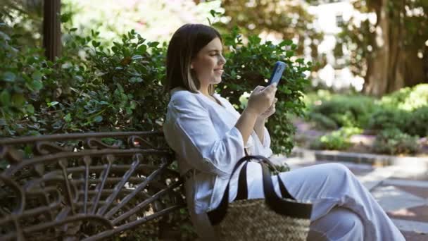 年轻美丽的惊慌失措的女人笑着坐在长椅上 在公园里用智能手机 — 图库视频影像