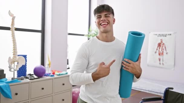 年轻的男子拿着瑜伽垫开心地微笑着 手指头指向康复诊所 — 图库视频影像
