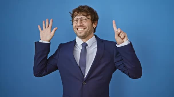 ビジネスススーツを着た若者は 自信と幸せを笑いながら指のナンバー6を指摘しています 単離された青い背景 — ストック動画